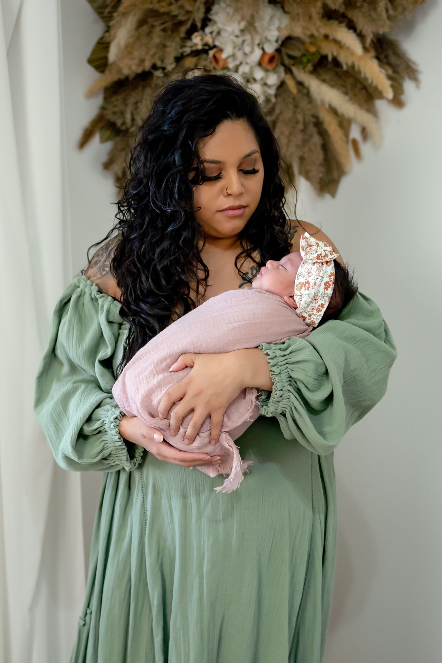 A person in a sage green dress cradles a baby wrapped in pink against a white backdrop.
