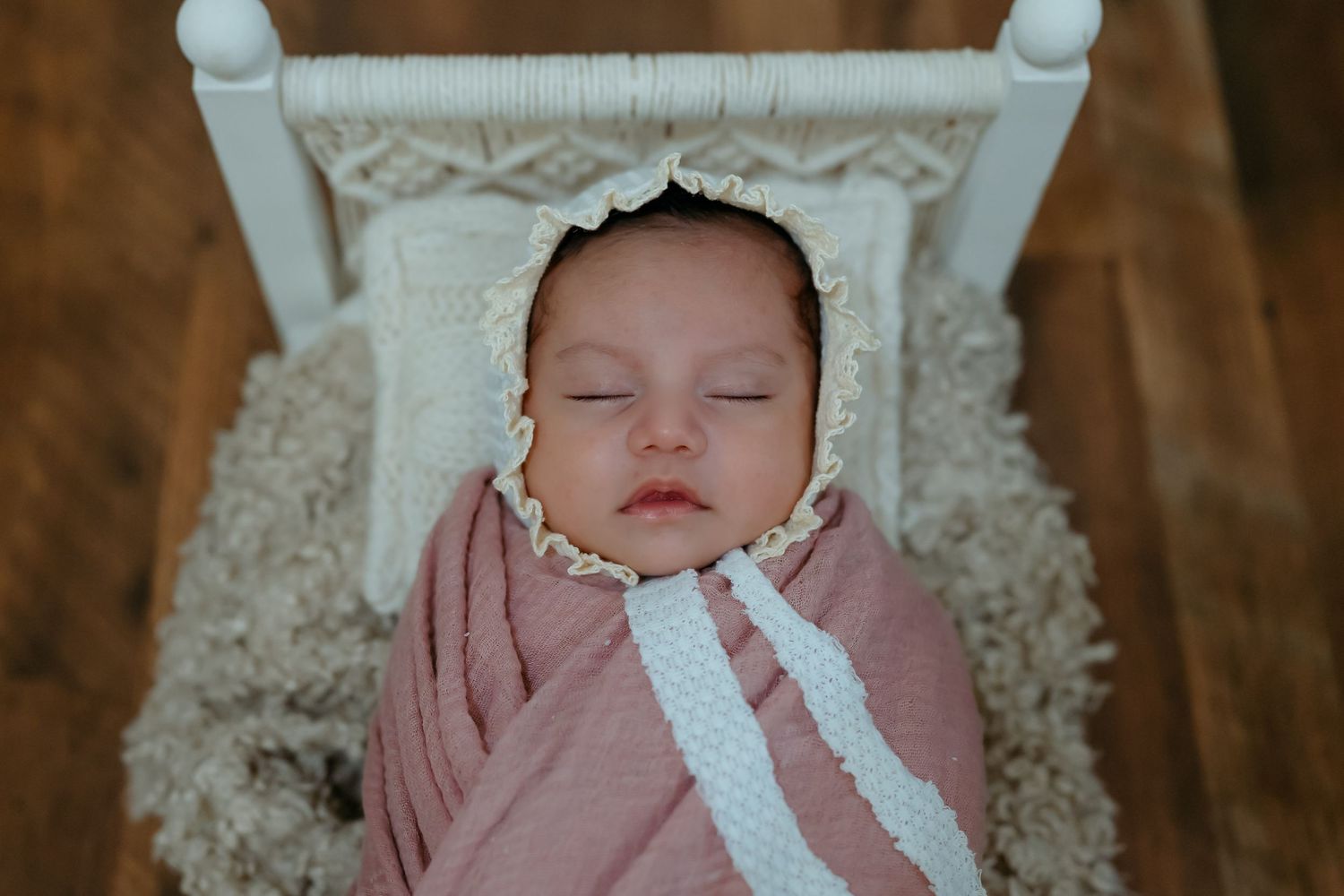 A sleeping newborn baby wearing a pink outfit and white lace bonnet rests peacefully on a textured blanket.