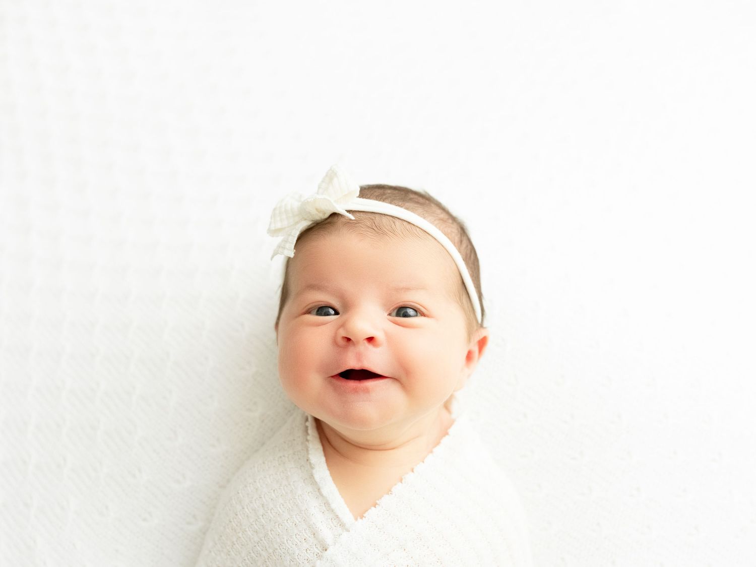 Happy baby wearing white headband and sweater smiles against white background.
