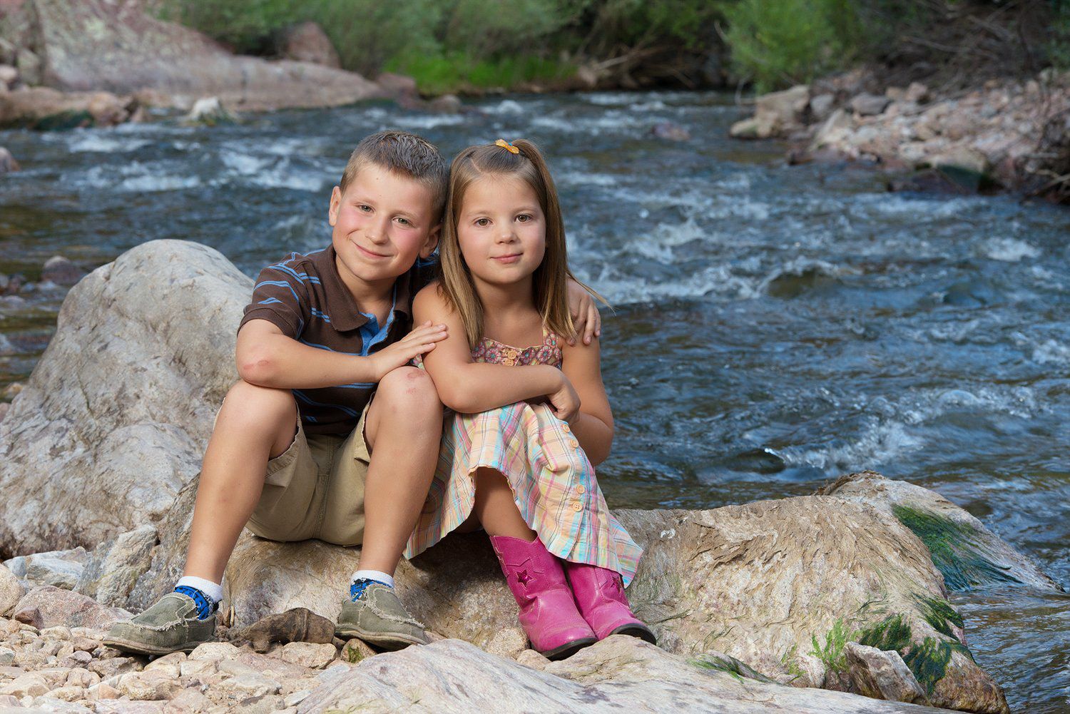 Two Erie Colorado children sit together on large rocks beside a rushing mountain stream in a scenic outdoor setting.