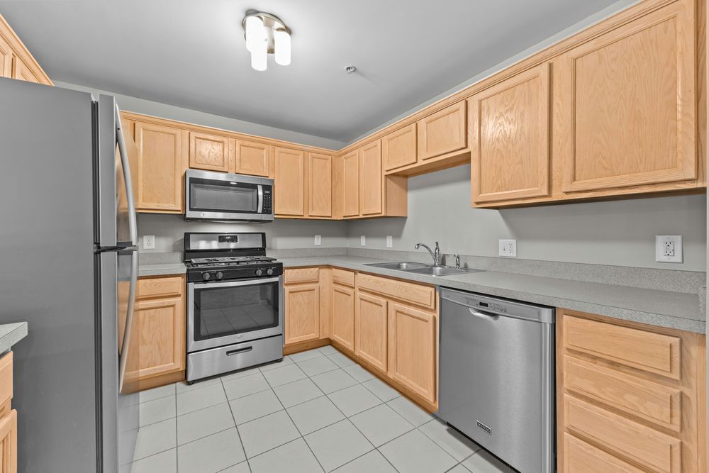 Modern kitchen with light wood cabinets, stainless steel appliances, tile floors, and a double sink.