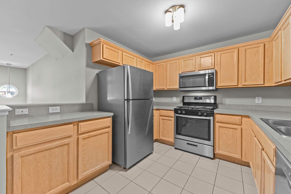 Kitchen with light oak cabinetry, stainless steel refrigerator, gas range, microwave, and tile flooring.