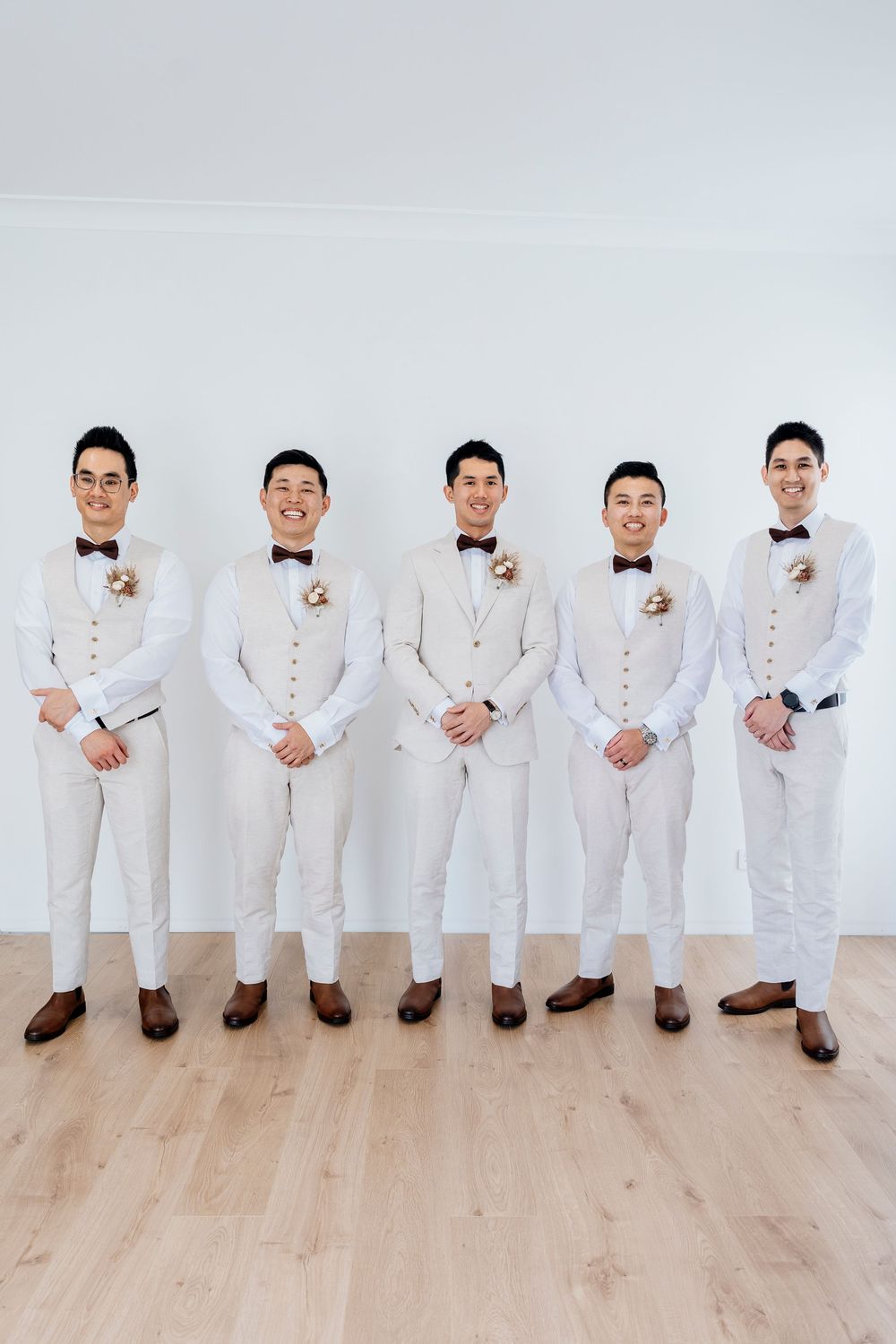 Groomsmen in matching white suits and bow ties pose together in a row against a light wooden floor.
