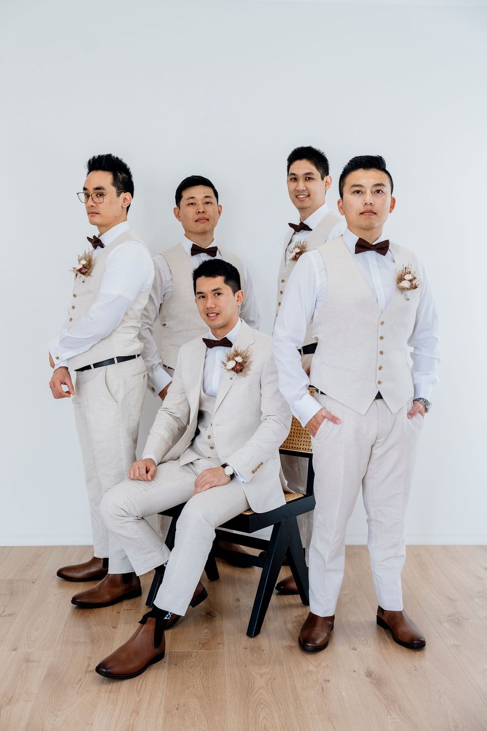Groomsmen in matching light gray suits and bow ties pose together against white wall with brown leather shoes.