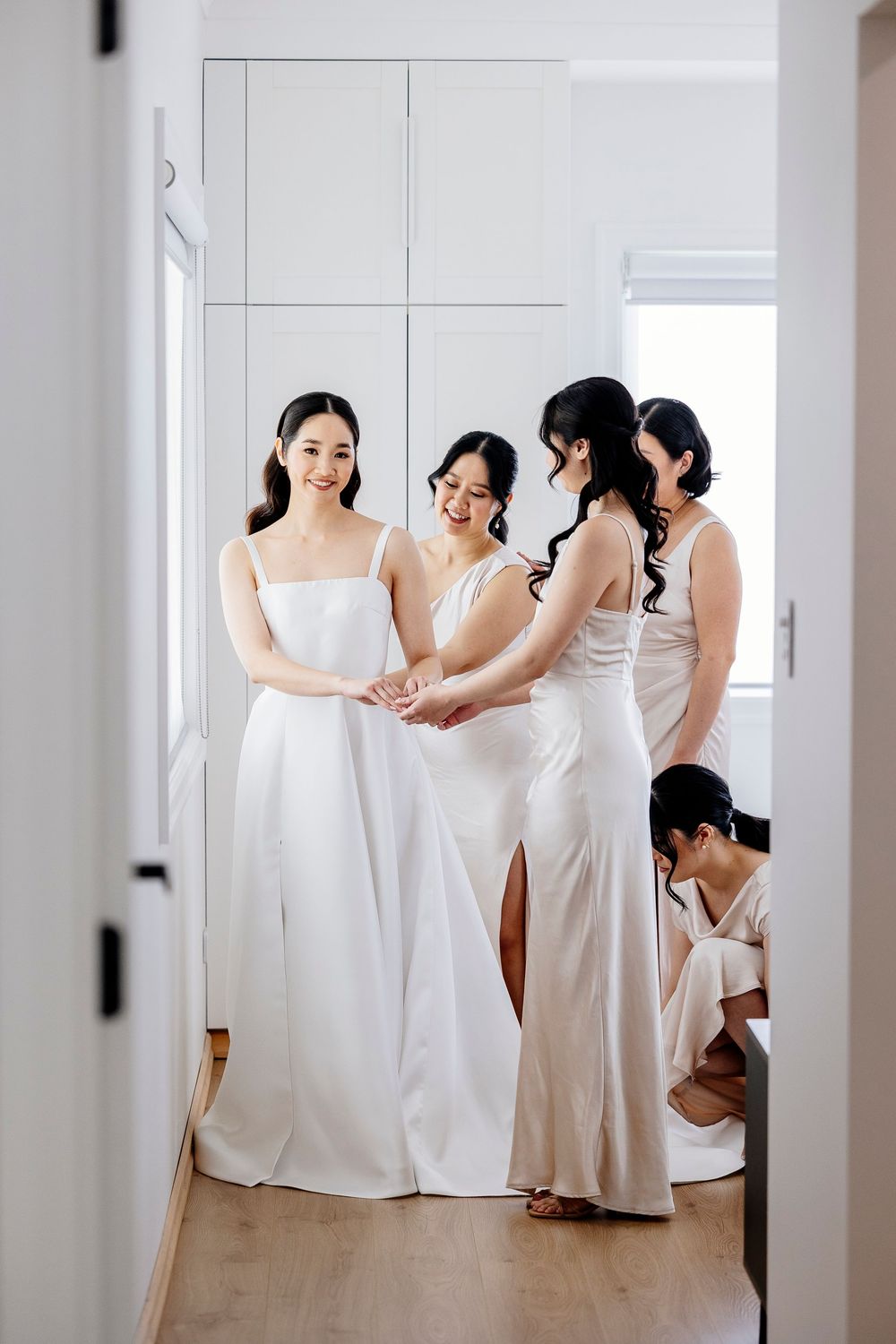 Bridesmaids in elegant white dresses gather and help each other prepare in a bright, minimalist room.