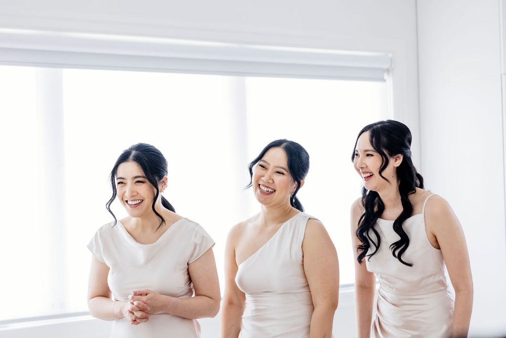 A group of women in white dresses sit together on a couch, laughing and smiling while sharing a joyful moment.