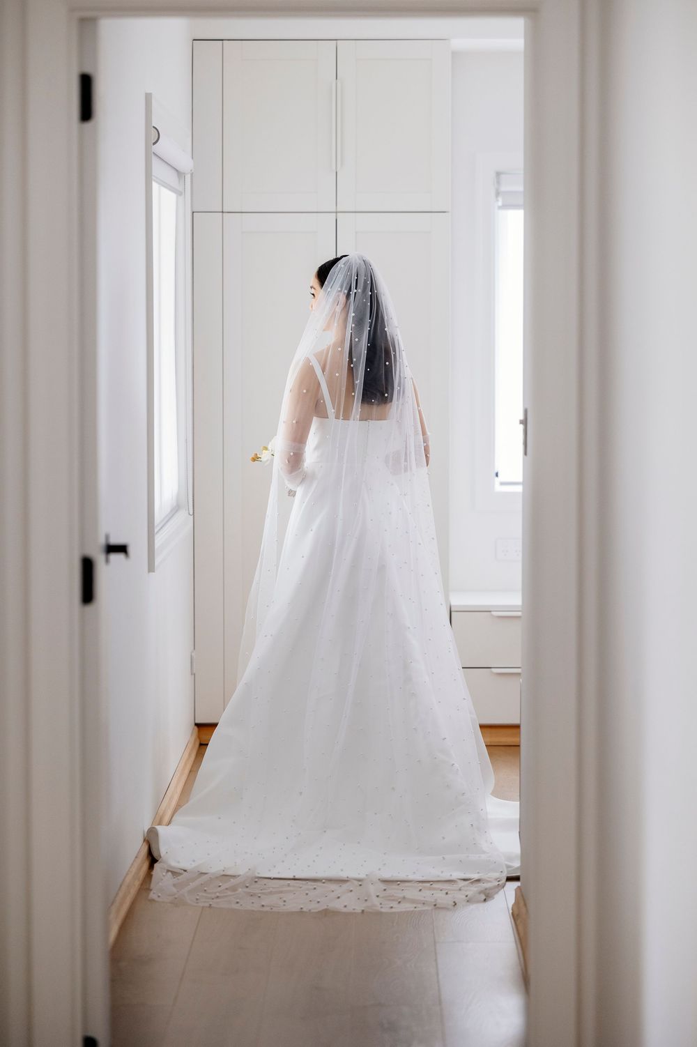 A bride in a flowing white wedding gown and long veil stands in a bright hallway, photographed from behind.