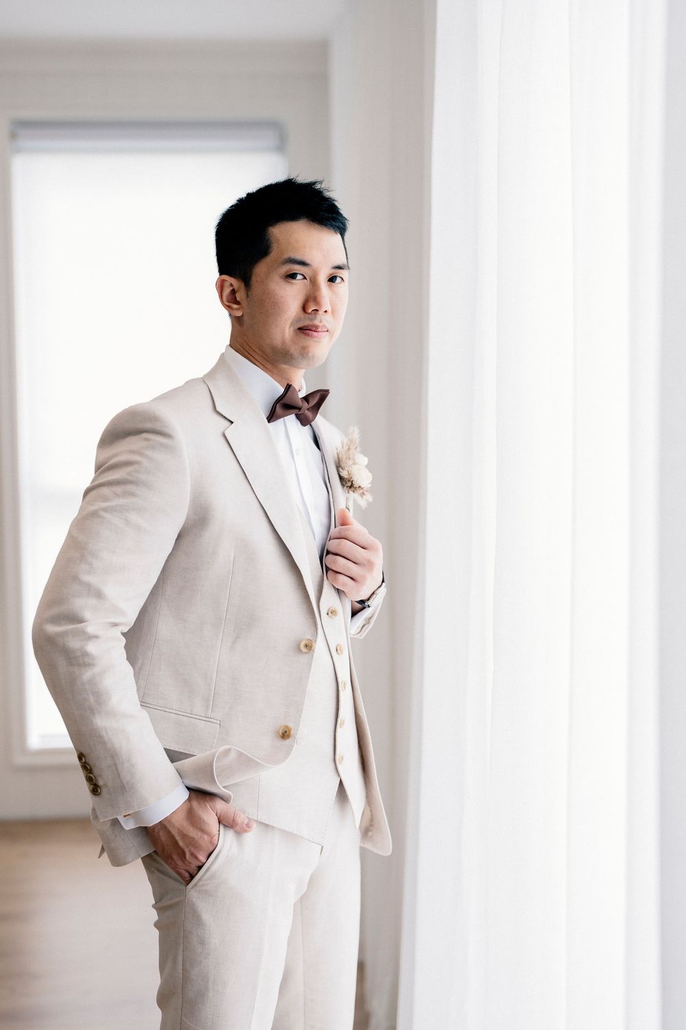 A groom in a crisp white suit poses elegantly in a bright room with natural window light.