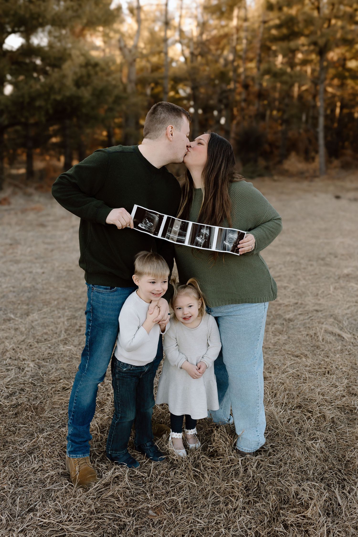 Family of four poses together in a golden field at sunset while holding ultrasound photos.