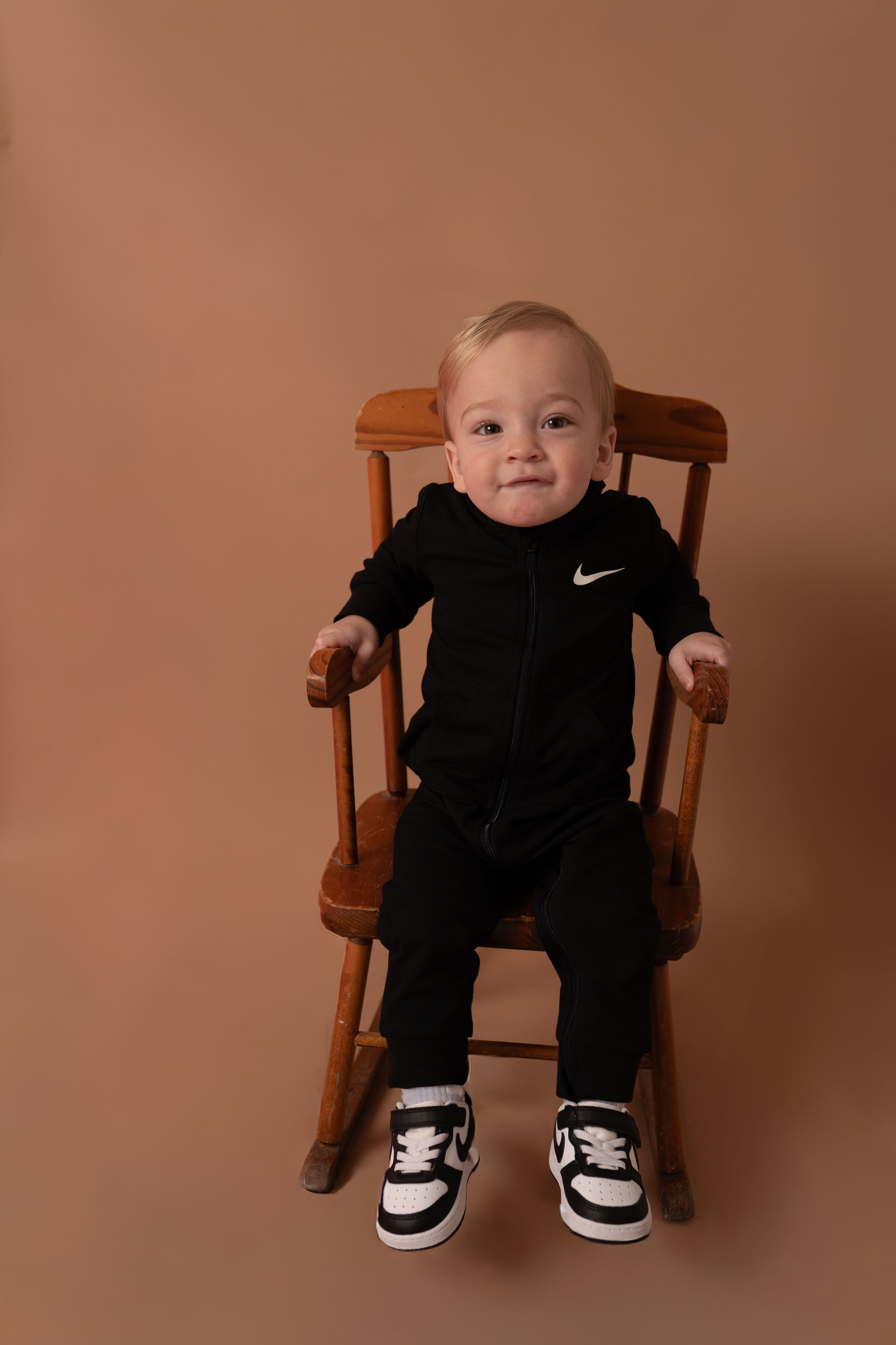 Baby wearing black Nike outfit sits in wooden chair against peach backdrop.