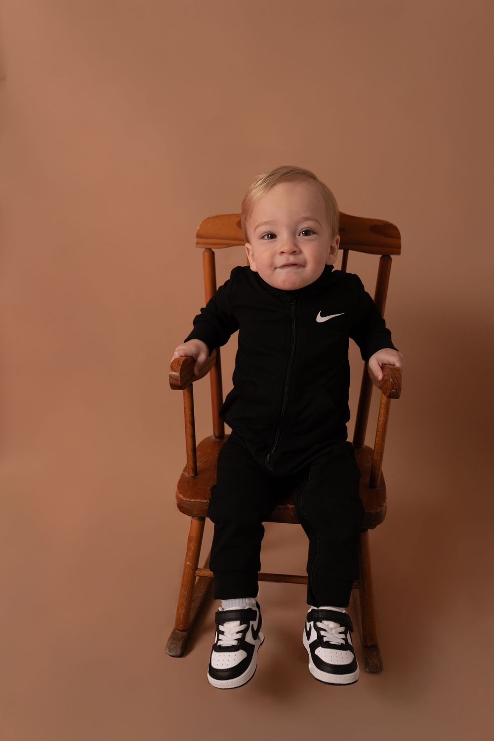 Baby wearing black Nike outfit sits in wooden chair against peach backdrop.