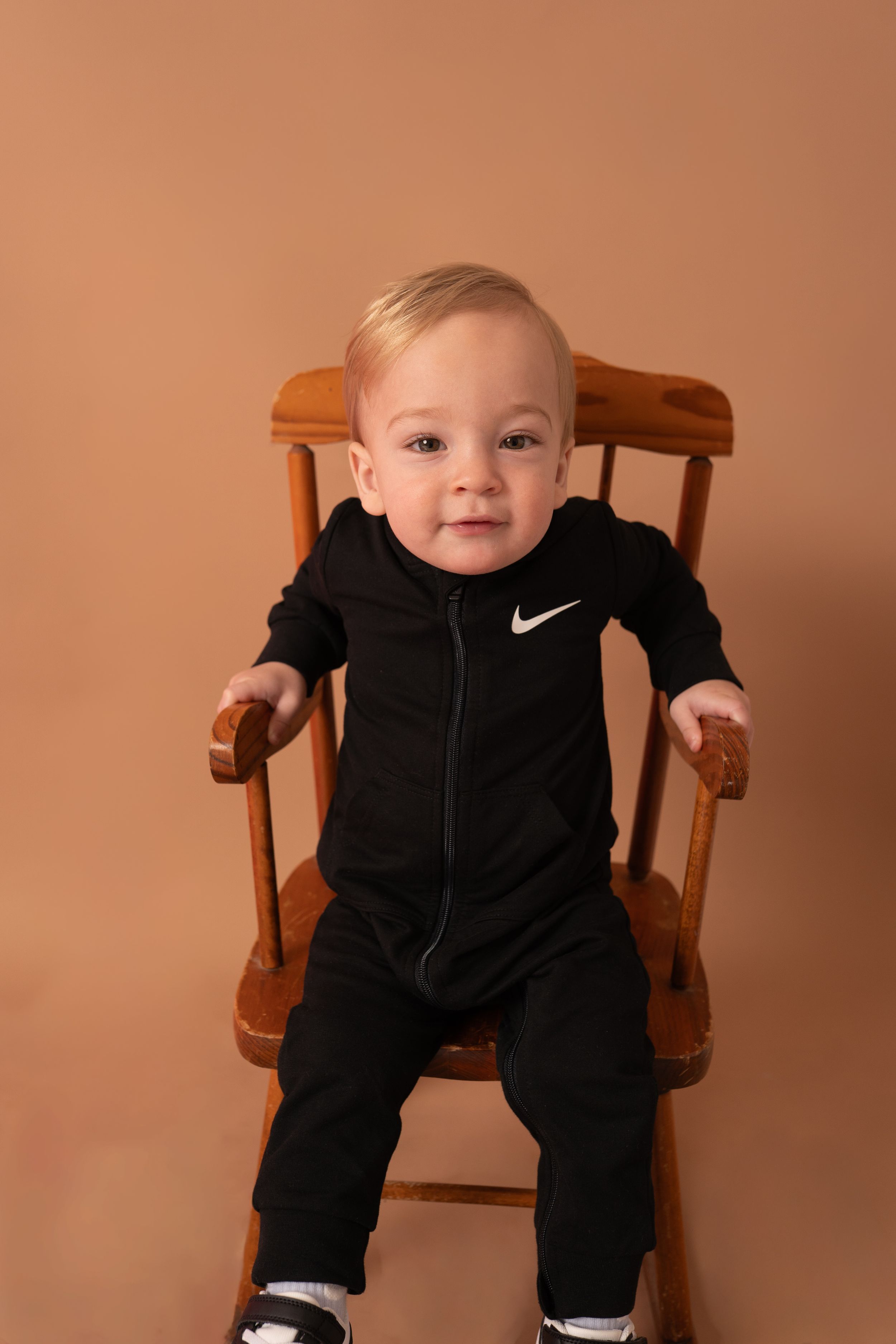 Happy baby in black Nike outfit poses in wooden chair.