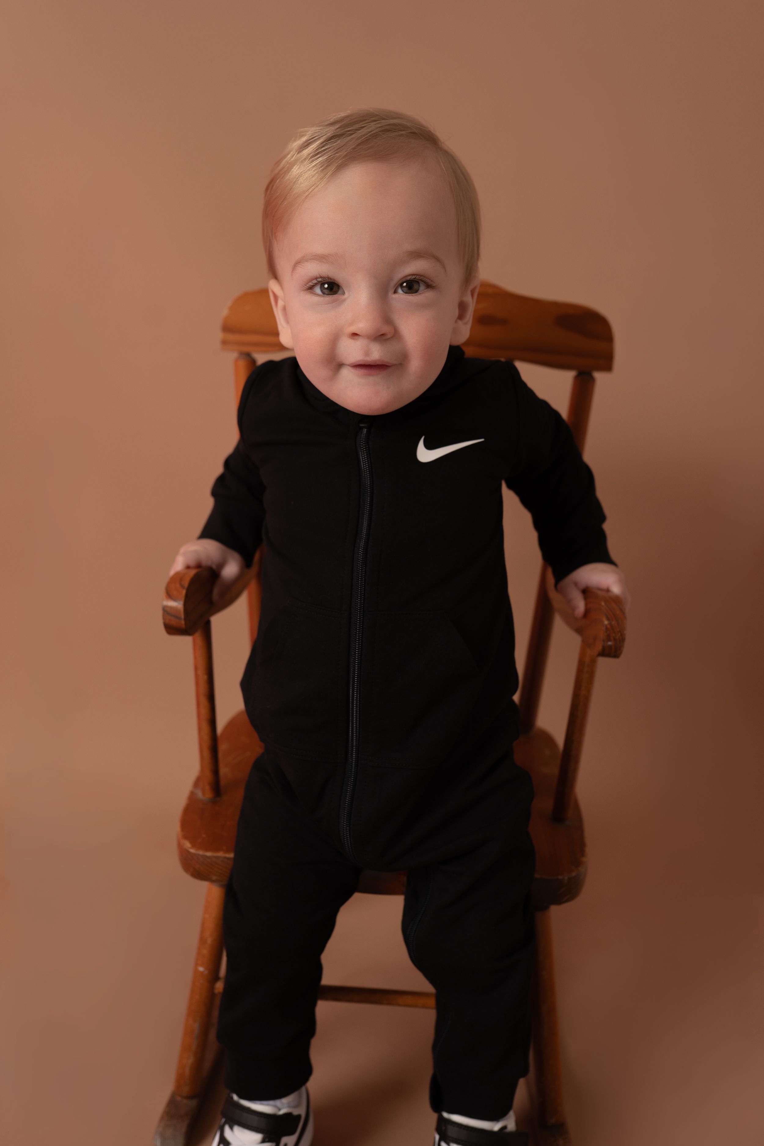 Smiling infant in black Nike tracksuit seated in wooden chair.