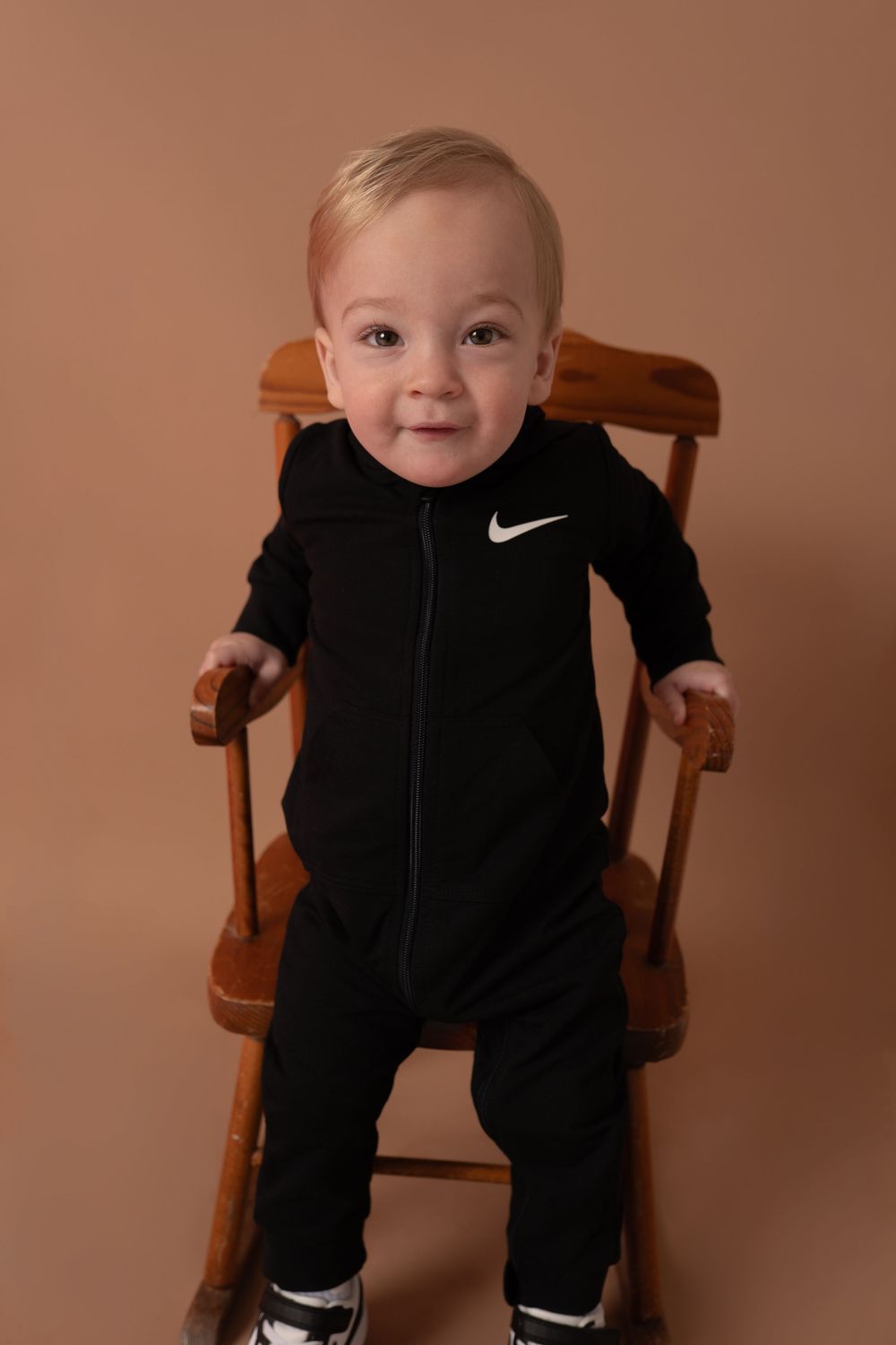 Smiling infant in black Nike tracksuit seated in wooden chair.