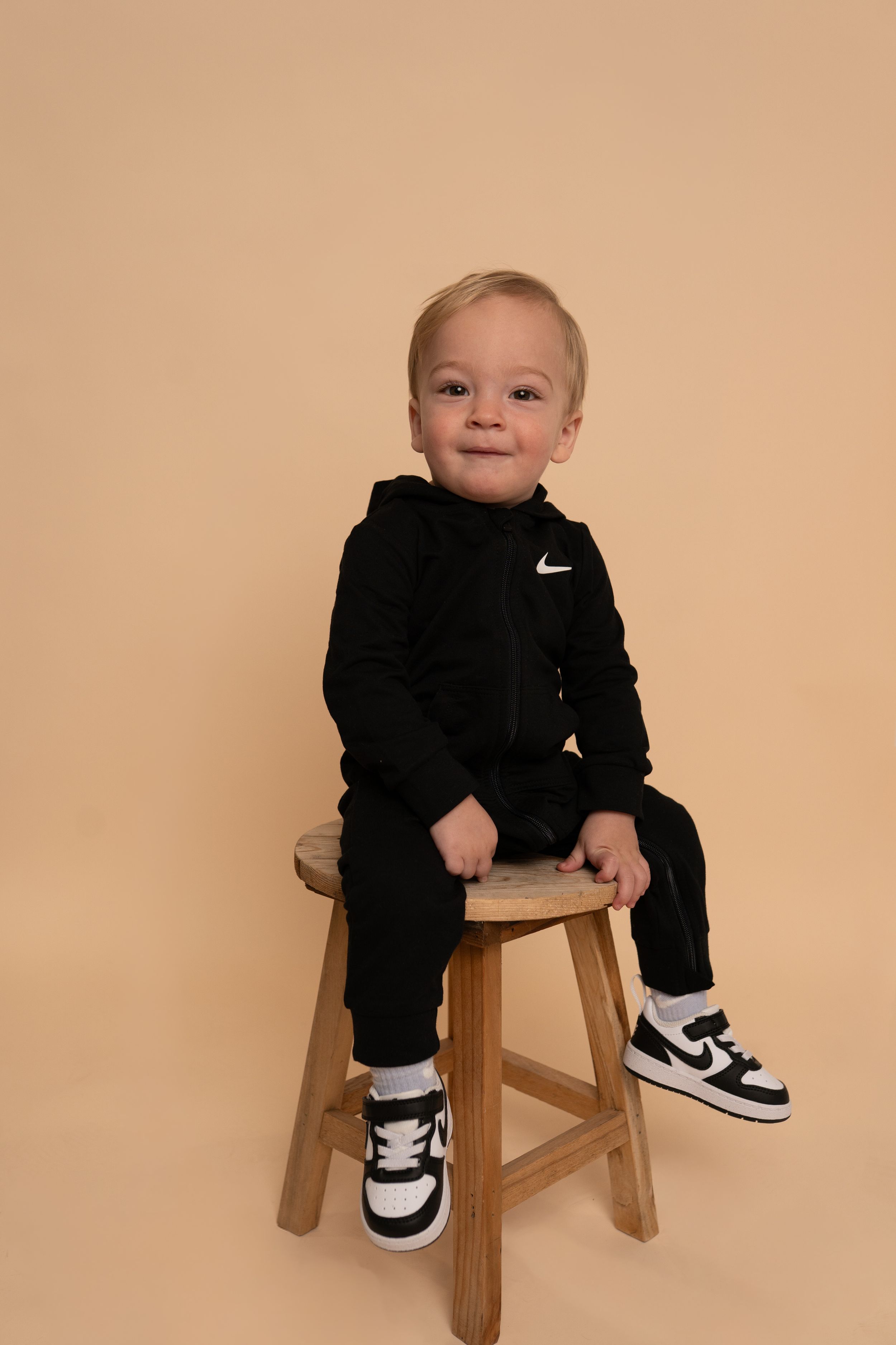 Playful baby wearing Nike outfit and sneakers sits in wooden chair.