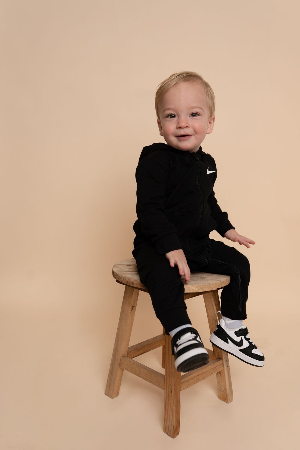 Cheerful infant in black Nike outfit gestures while sitting in wooden chair.