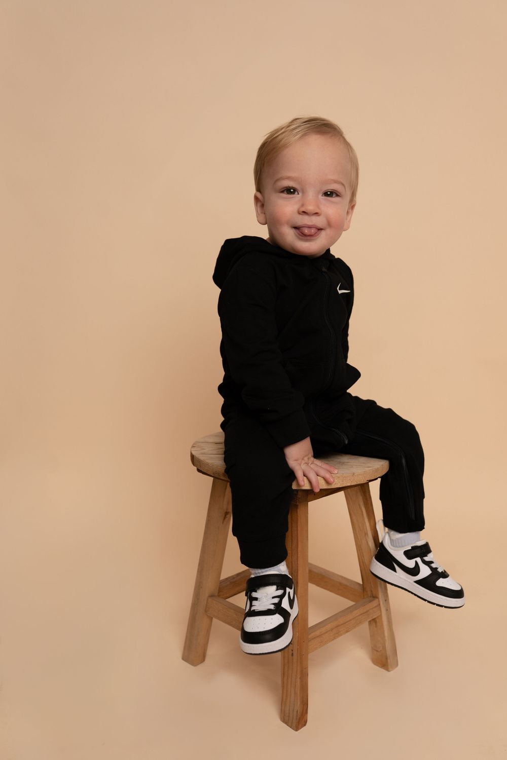 Baby in Nike tracksuit and sneakers poses playfully on wooden chair.