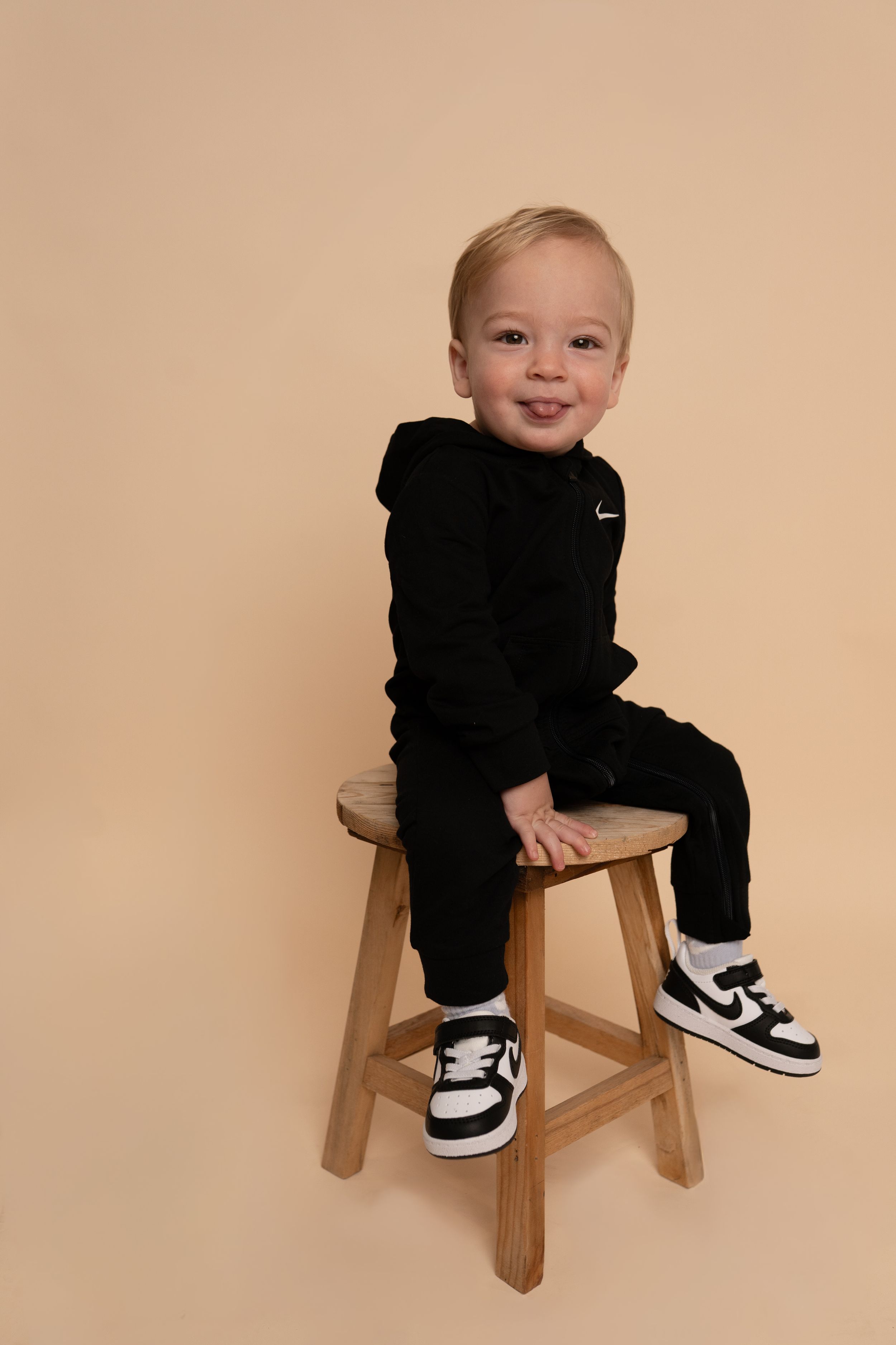 Baby in Nike tracksuit and sneakers poses playfully on wooden chair.
