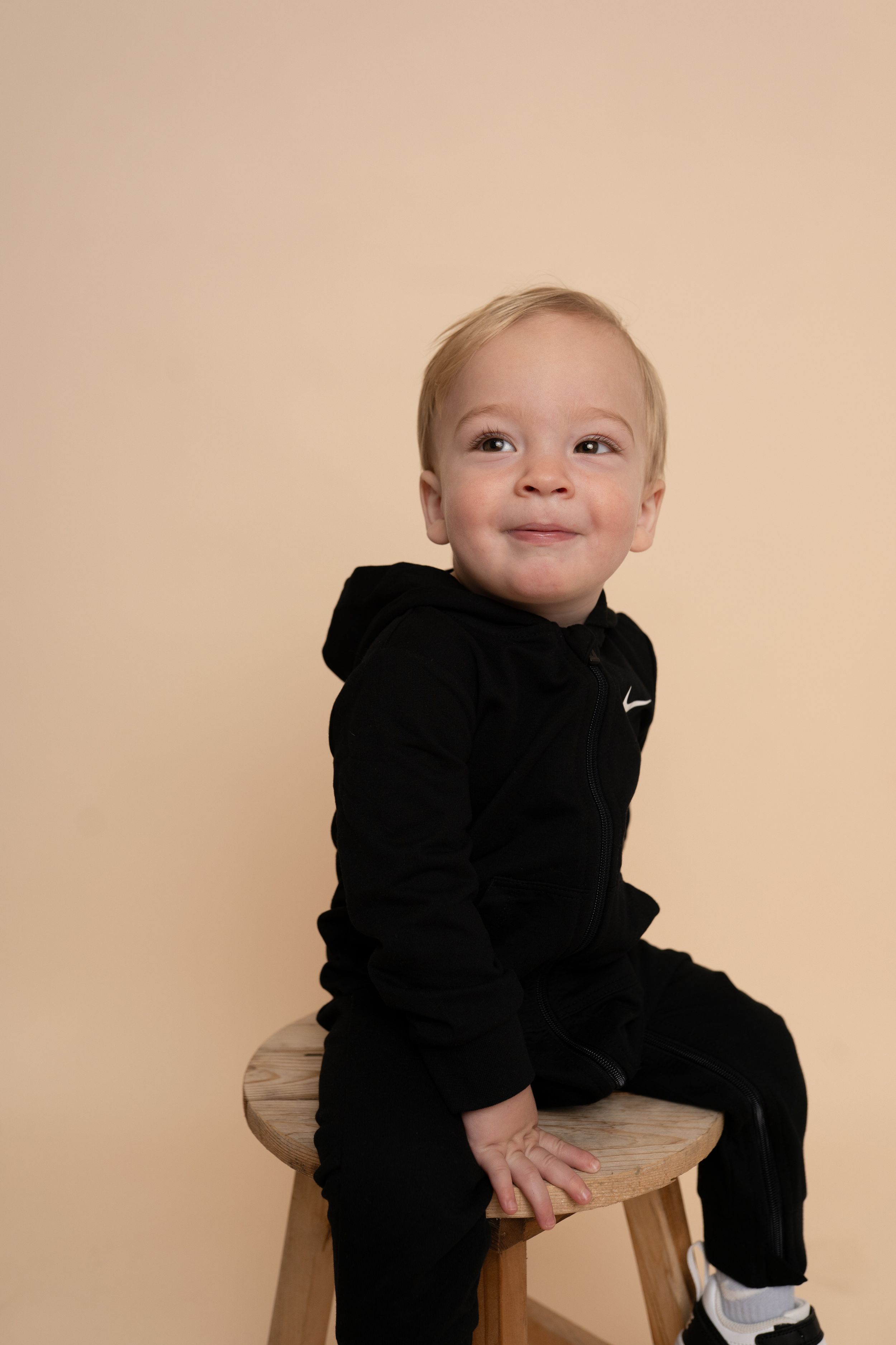 A small child wearing a black Nike sweatsuit sits on a wooden stool against a beige backdrop.