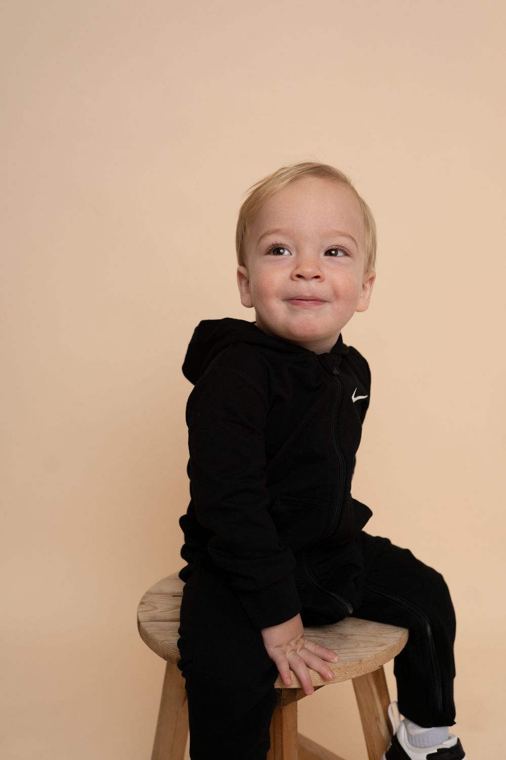 A small child wearing a black Nike sweatsuit sits on a wooden stool against a beige backdrop.