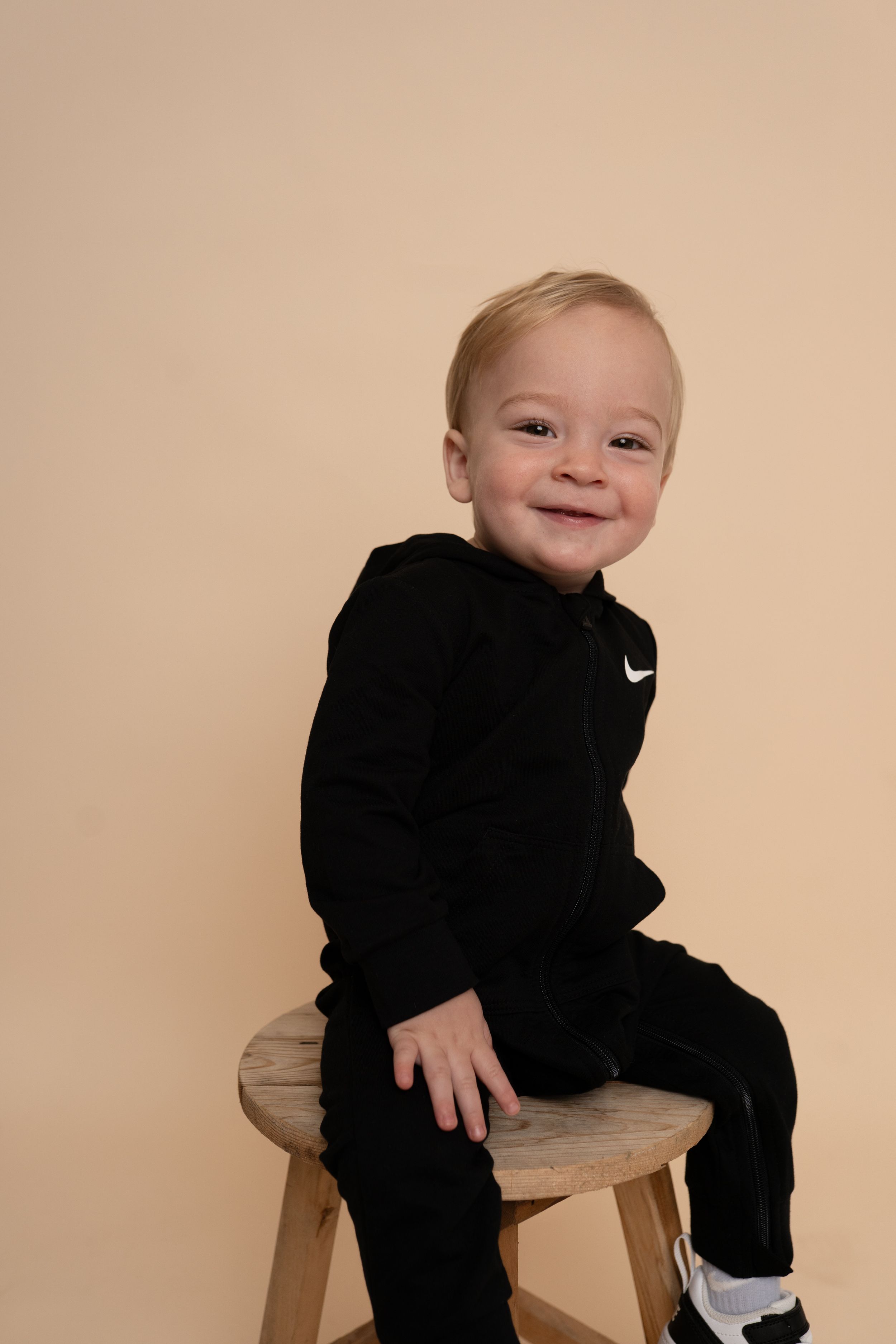 Toddler in black Nike outfit smiles while sitting on wooden stool against neutral background.