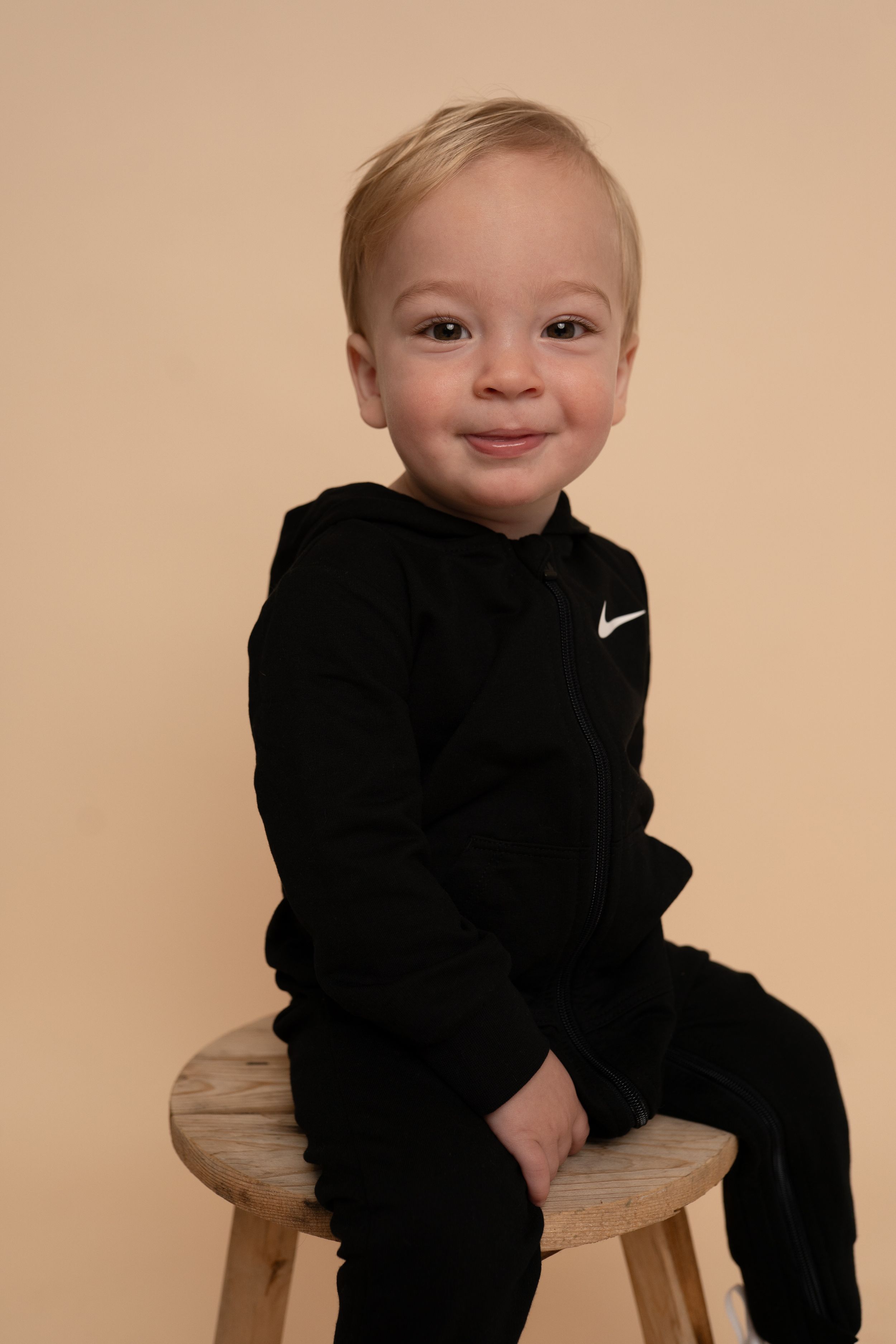 Young child in black hoodie poses playfully on wooden stool during studio photoshoot.