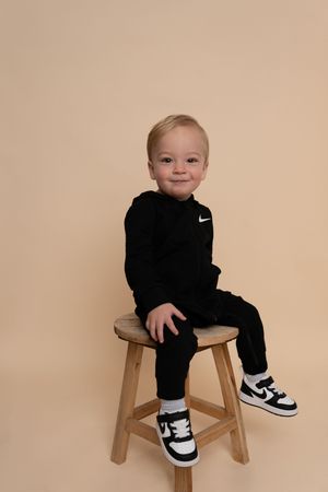 Small child in black Nike outfit and sneakers sits casually on wooden stool.