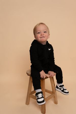 Toddler wearing black sweatsuit and white sneakers poses on wooden stool in studio setting.