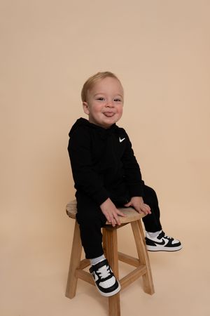 Young child in black Nike outfit smiles while perched on wooden stool against beige background.