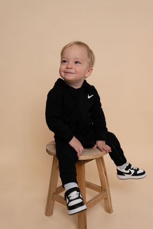 Small child in black Nike sweatsuit sits playfully on wooden stool during photo session.