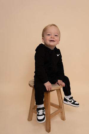 Toddler wearing black Nike outfit and sneakers poses on wooden stool in studio.