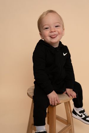 Young child in black Nike sweatsuit smiles while sitting on wooden stool during photoshoot.