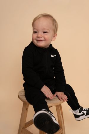 Small child in black Nike outfit laughs while posing on wooden stool against neutral backdrop.