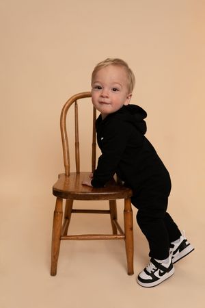Baby wearing black clothing and sneakers interacting with a wooden chair in studio setting.
