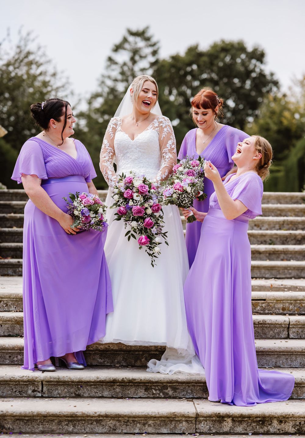 Bridesmaids in flowing purple dresses share candid laughing moments with bride in white on stone steps during photoshoot.