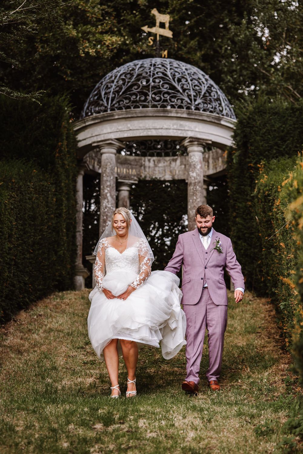 A couple in wedding attire walks together through a garden path under an ornate metal archway. Relaxed Dorset Wedding photographer