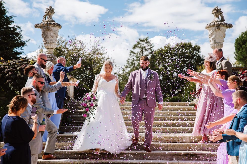 Groom in purple suit Wedding guests toss pink and purple flower petals at a couple descending stone steps in a garden celebration.