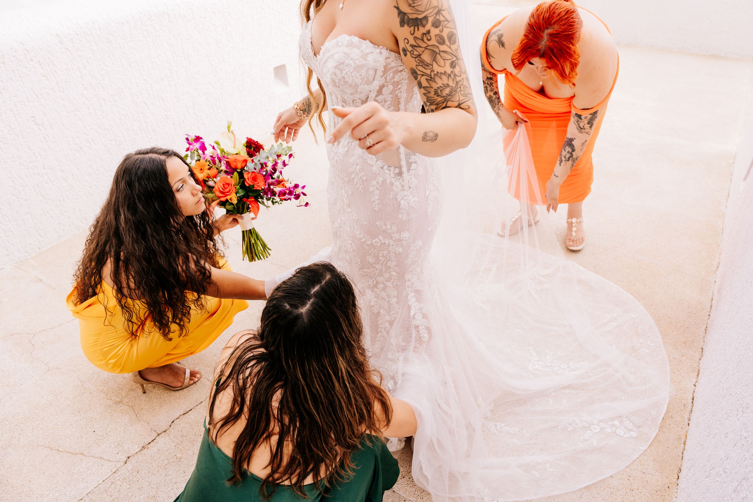 Bridesmaids helping bride in lace wedding gown, one holding colorful bouquet, another adjusting dress train outdoors.