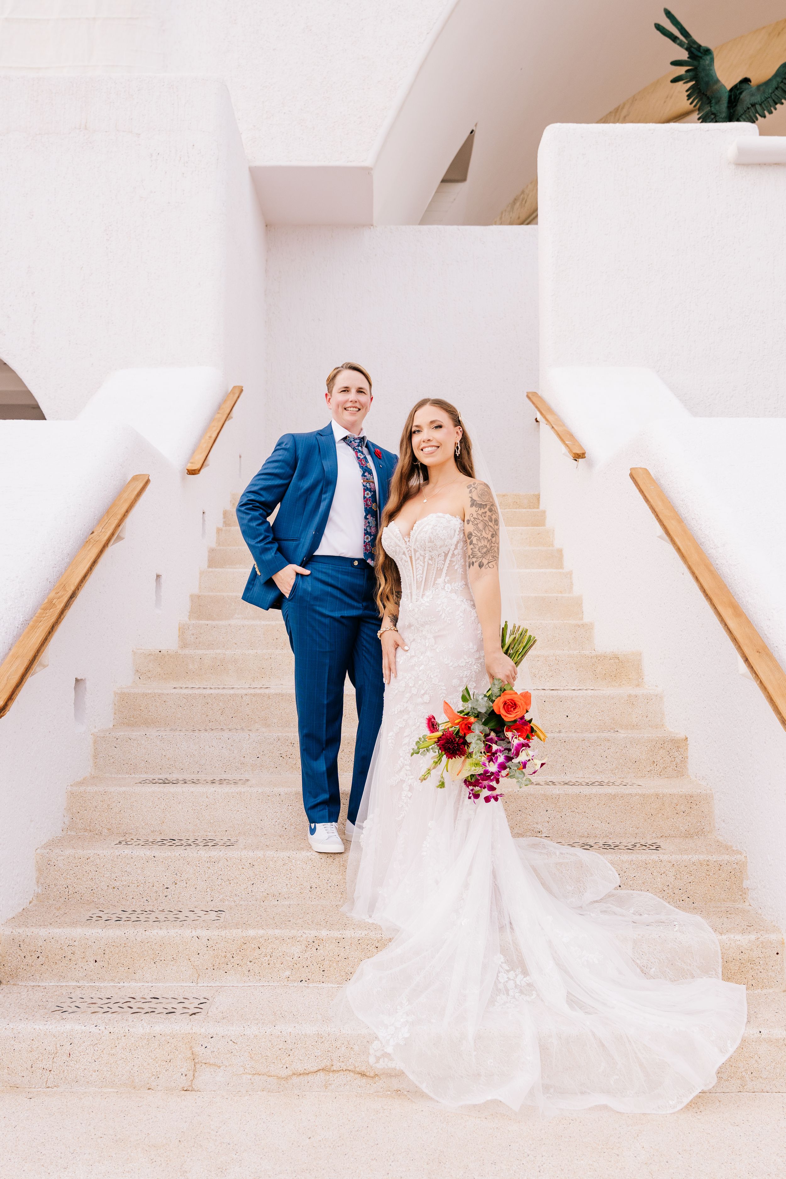 Bride in white gown holding colorful bouquet stands with groom in blue suit on elegant white staircase.
