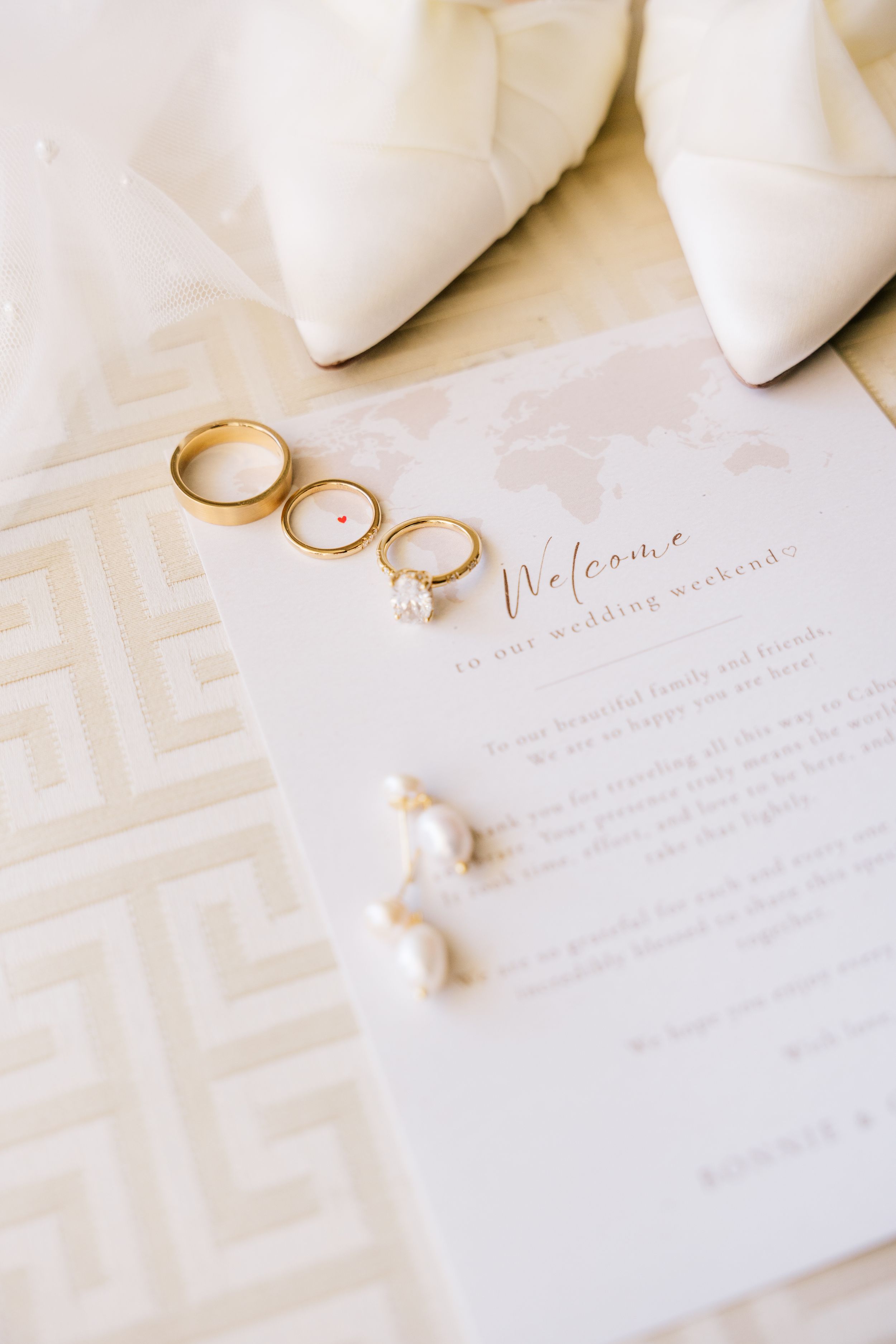 Gold wedding bands and diamond engagement ring beside pearl earrings on a wedding weekend welcome card with white heels.