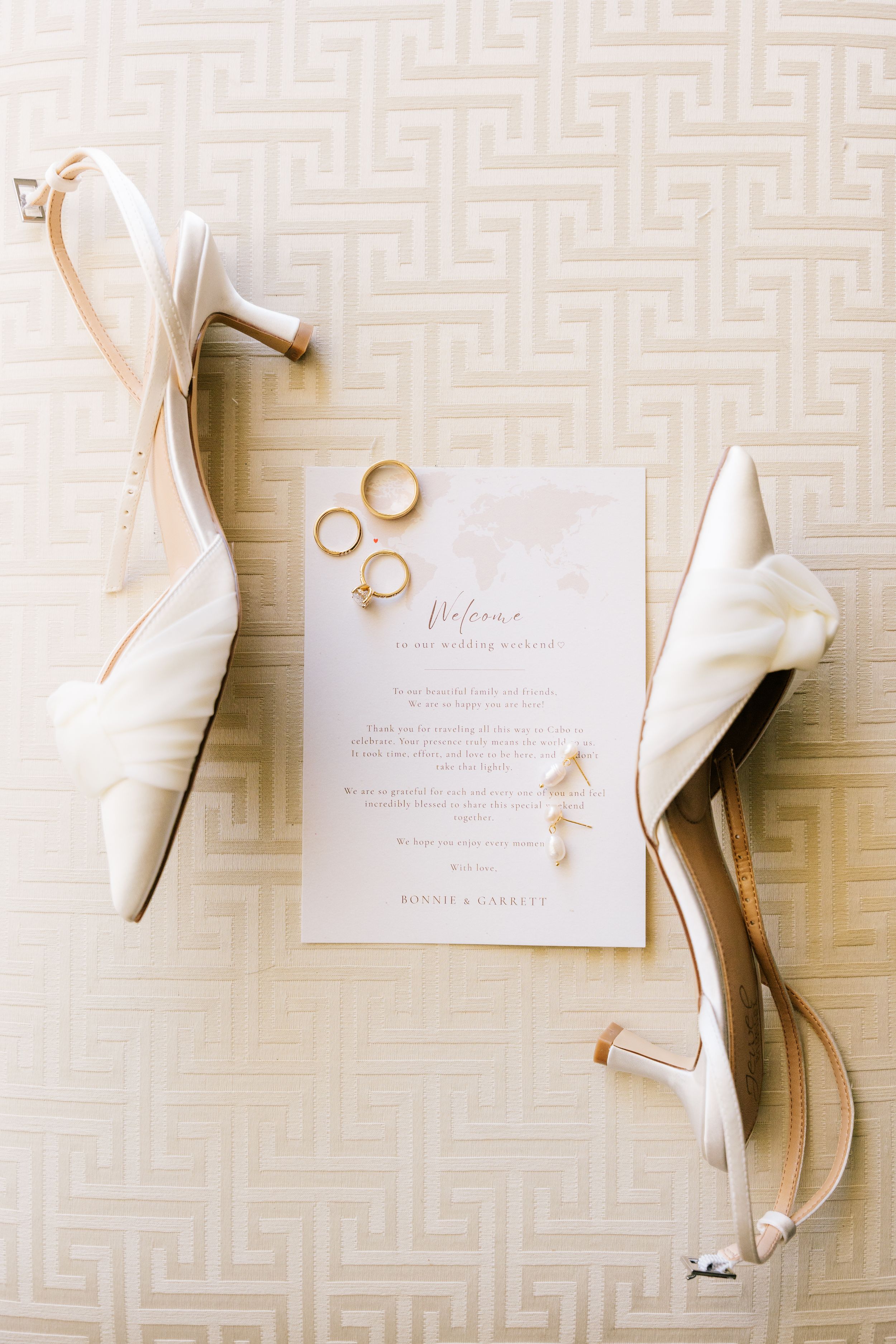 White satin wedding heels beside gold rings and an elegant wedding invitation on a cream geometric patterned surface.