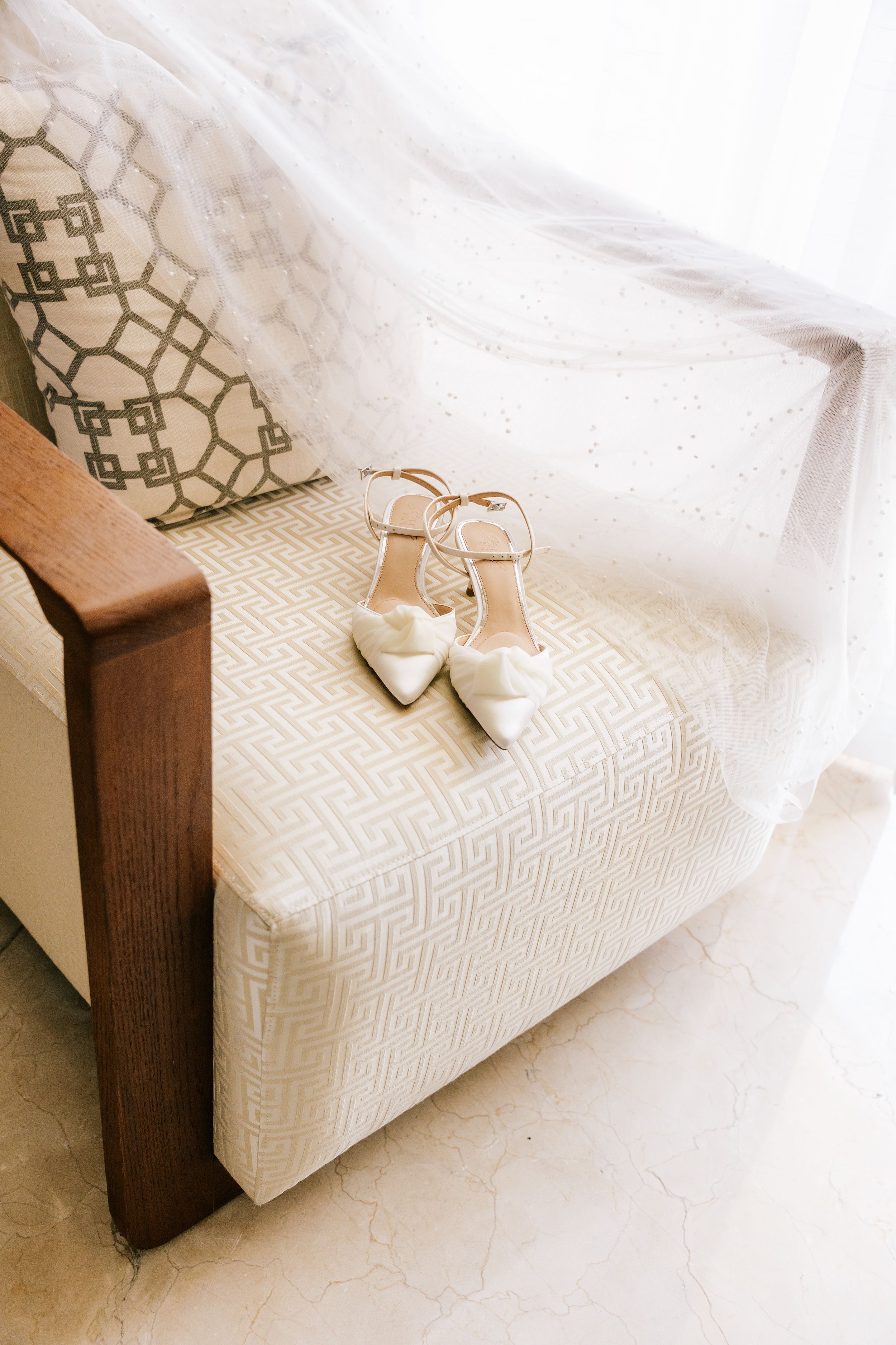 Elegant white bridal heels with ankle straps resting on a cream patterned armchair with a flowing veil.