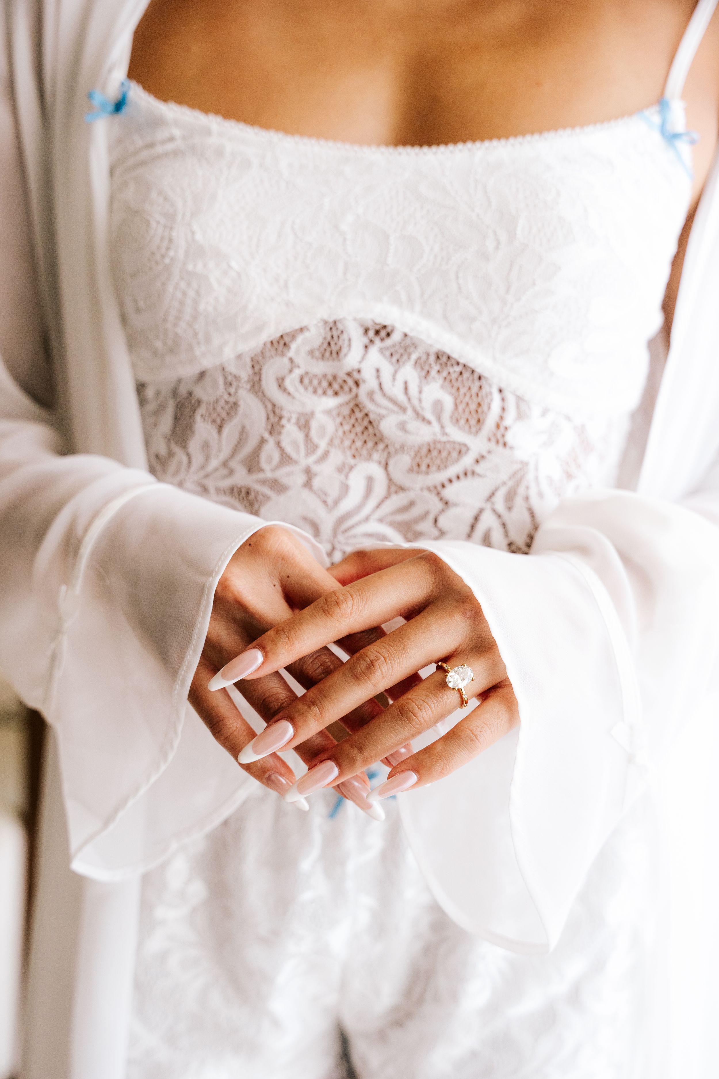 Bride displaying elegant engagement ring with French manicure, wearing white lace bridal outfit and sheer robe.