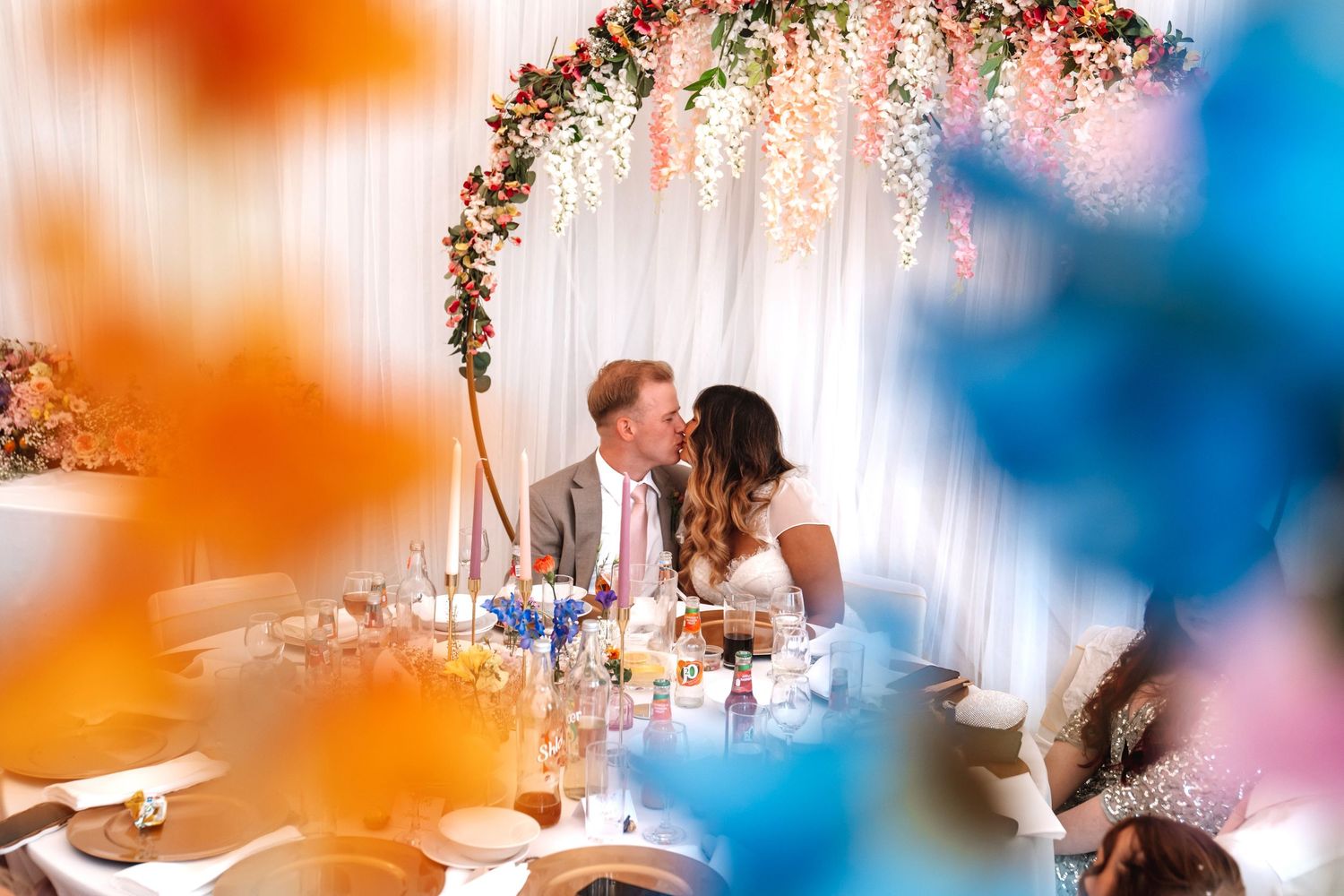 Couple sharing a kiss at their wedding reception table adorned with floral arch and colorful balloon decorations.