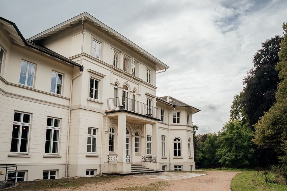 Panoramic view of a white historic mansion with multiple wings and tall windows set against a cloudy sky.
