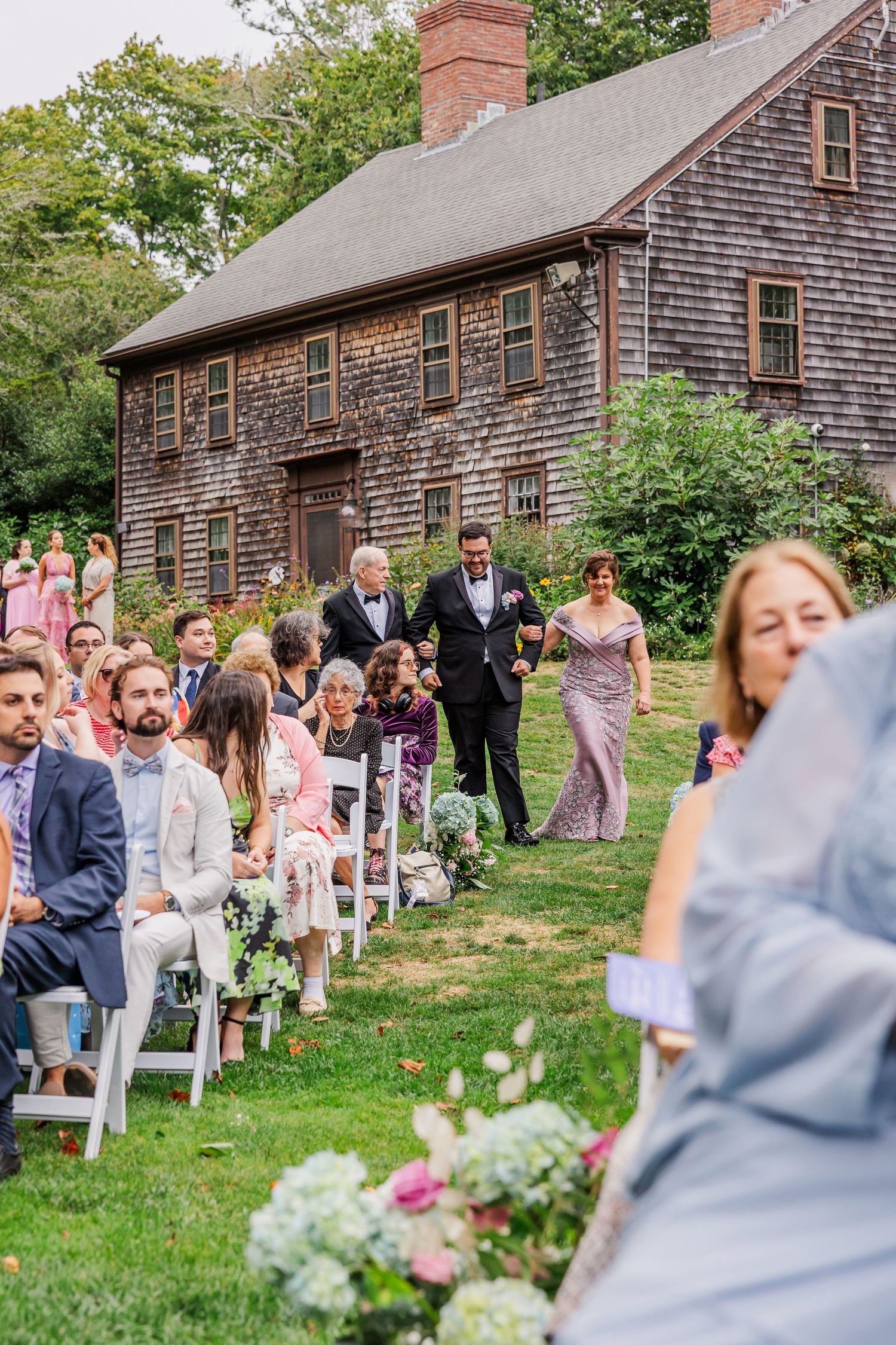 Rustic outdoor wedding ceremony setup with white chairs on grass lawn in front of historic brown wooden buildings.