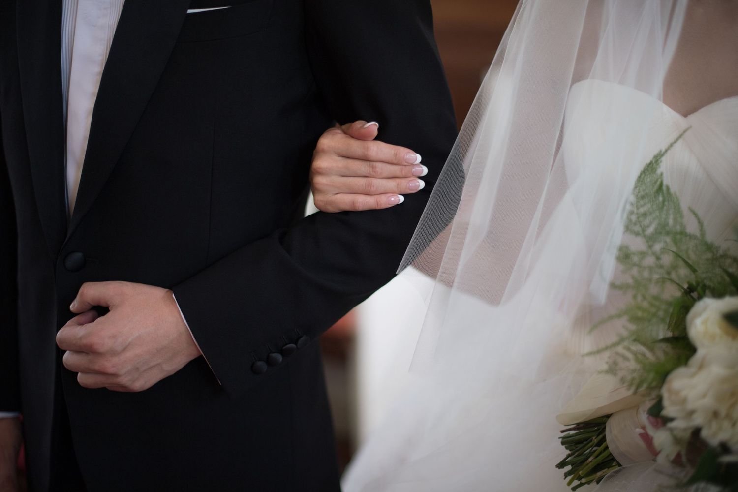 Bride and groom holding hands at wedding ceremony, bride holding white bouquet wearing veil, groom in black suit.