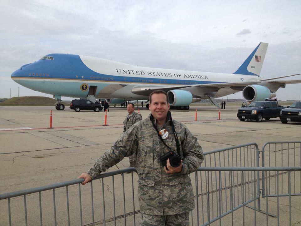 Air Force One, the presidential aircraft, parked on a tarmac with photographer Ashley Bell and barriers visible.