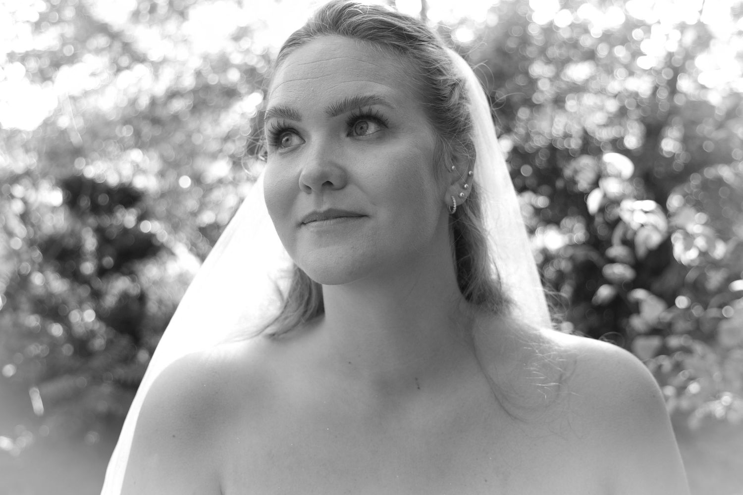 Black and white close-up portrait of bride wearing veil looking upward in a dreamy outdoor setting.