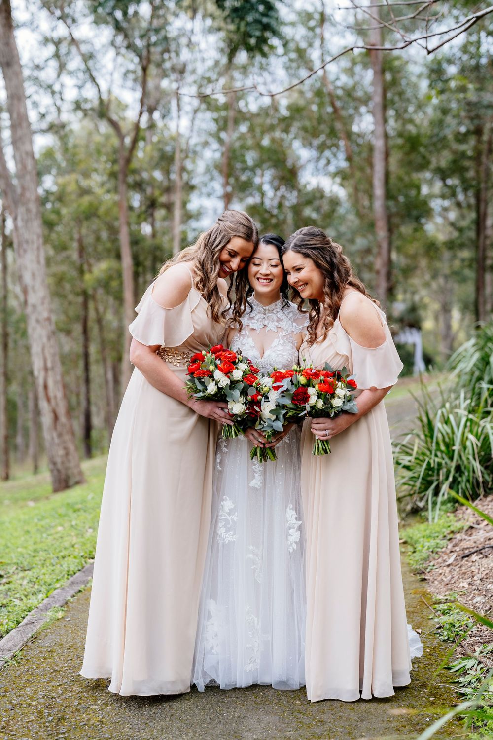 Bridesmaids in blush dresses share a candid laughing moment with the bride in a white gown at an outdoor woodland wedding.